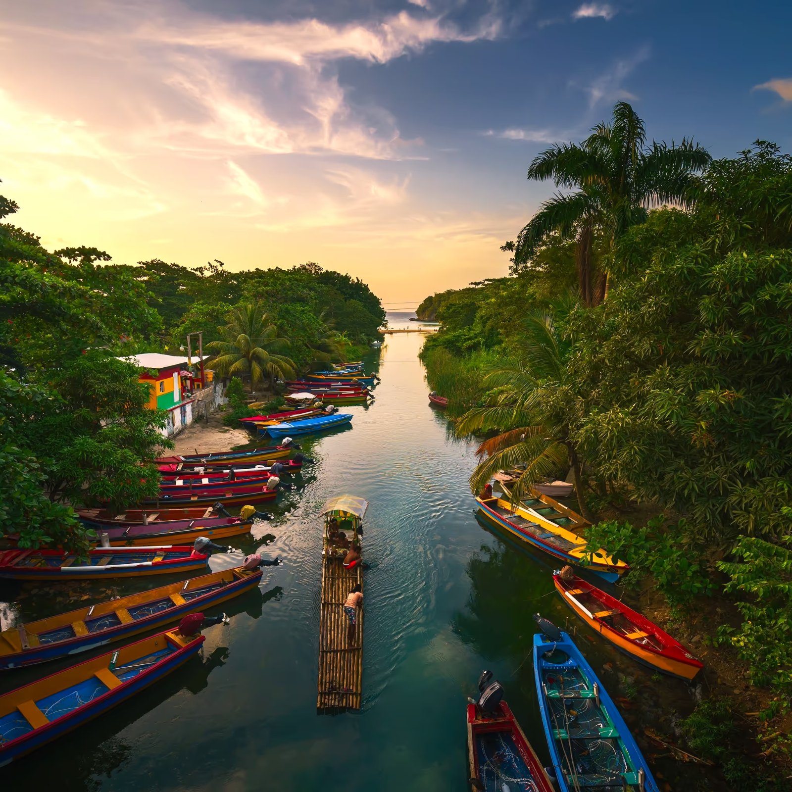 Bunte Holzboote und ein Bambusfloß auf dem White River bei Ocho Rios, Jamaika, umgeben von tropischem Grün und Palmen im Abendlicht